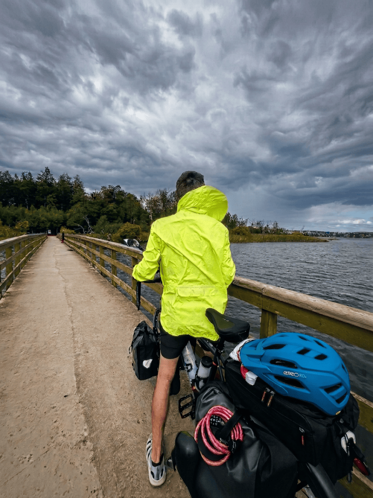 A person in a bright yellow rain jacket stands beside a bicycle with bags, overlooking a path by a body of water under a cloudy sky.