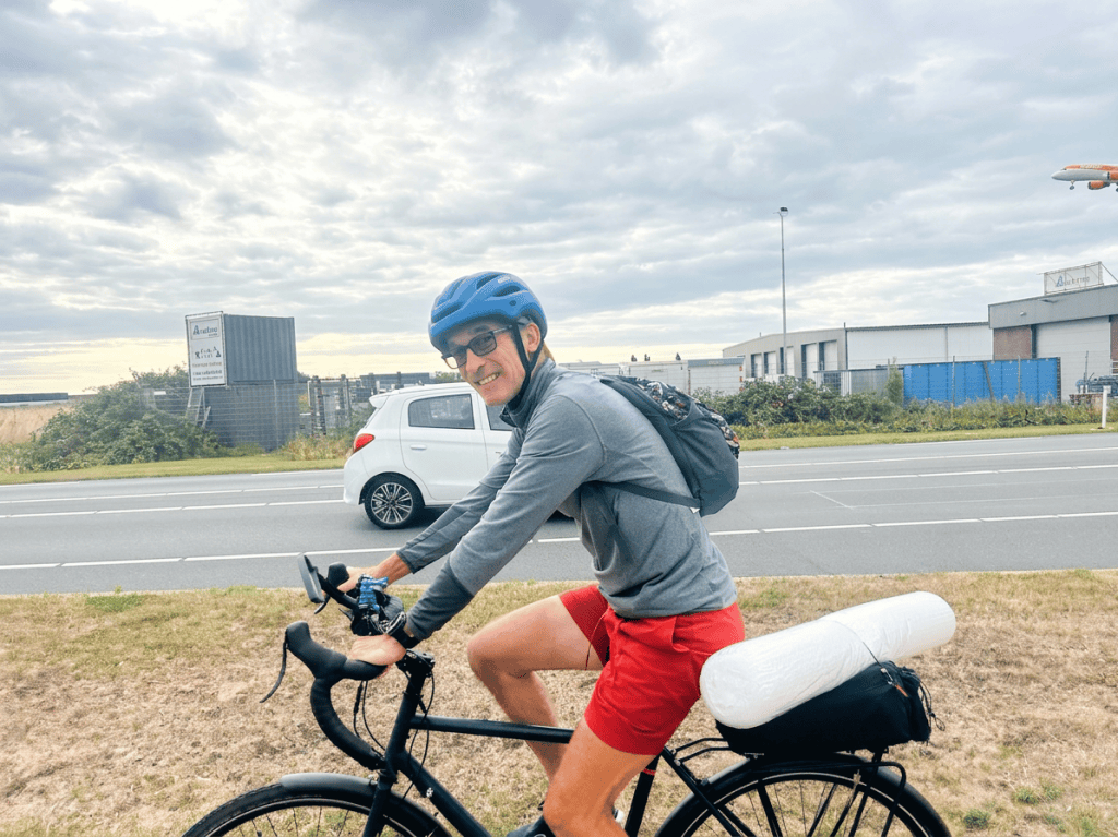 A cyclist wearing a blue helmet and sunglasses smiles while riding a black bicycle. The cyclist is dressed in a grey long-sleeve shirt and red shorts, with a backpack on their back. In the background, a white car is passing on the road, and an airplane is flying overhead against a cloudy sky.
