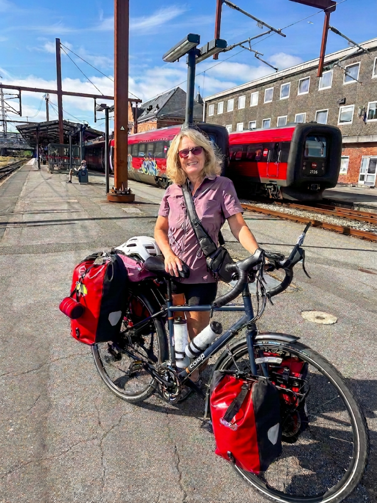 A woman with curly blonde hair stands next to a loaded bicycle at a train station, with red trains and a blue sky in the background.