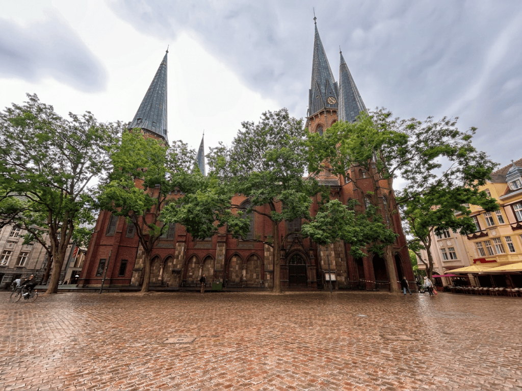 Historical red brick church with tall spires surrounded by trees and a cobblestone square under a cloudy sky.