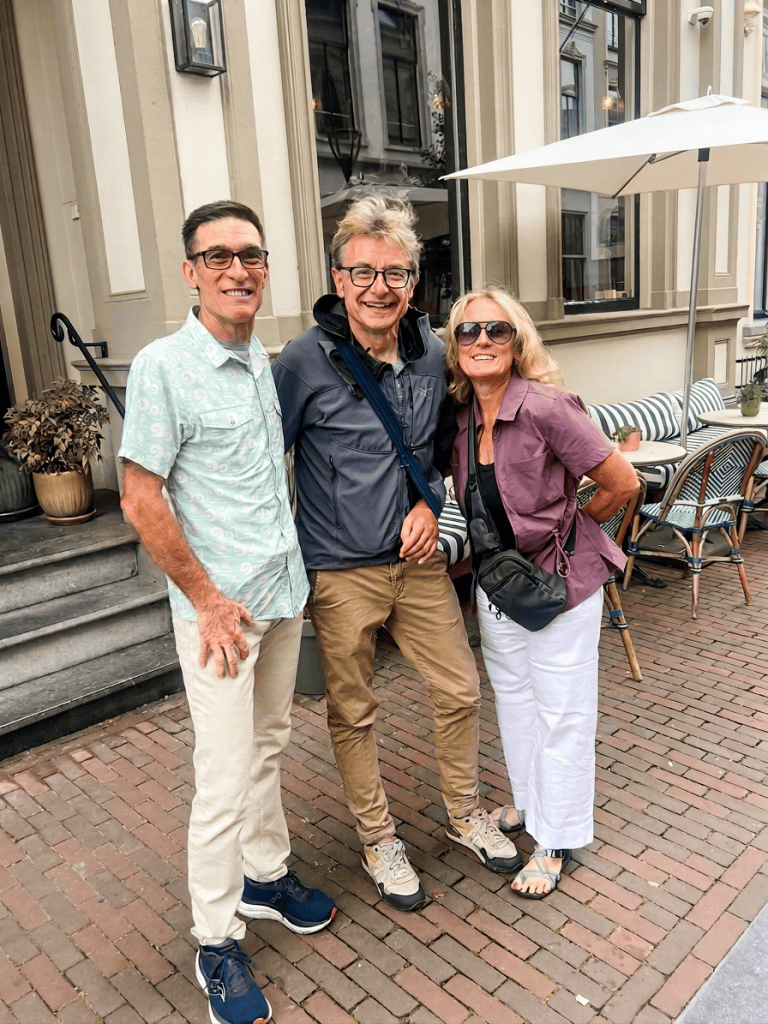 Three people smiling and posing outdoors in front of a café with a brick pathway and tables. The first person is wearing a light green shirt, the second is in a dark jacket, and the third is in a purple top.