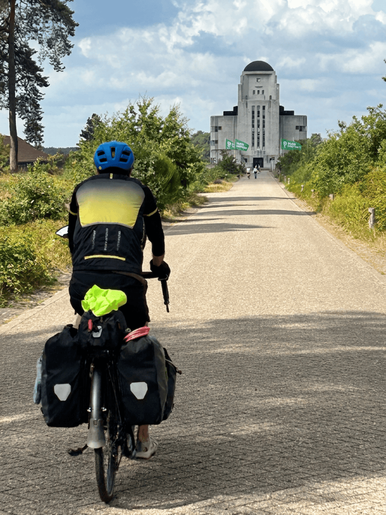 A cyclist with a blue helmet rides along a gravel path lined with greenery, approaching a large, modern architectural structure in the distance.